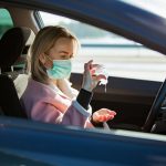 Woman in protective mask sitting in a car on road, using hand sanitizer.
