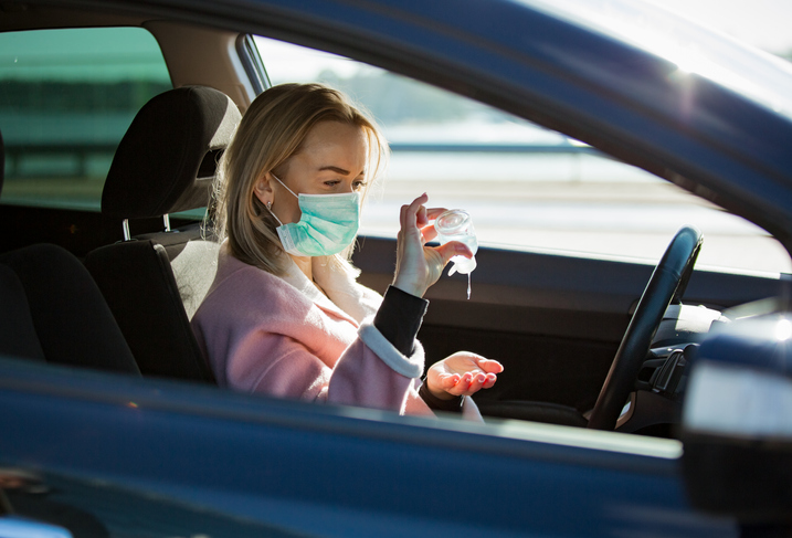 Woman in protective mask sitting in a car on road, using hand sanitizer.